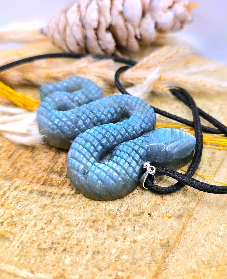 A coiled blue-green stone pendant necklace with a textured scale pattern, threaded on a black cord, displayed on a sandy surface with a pinecone in the background.