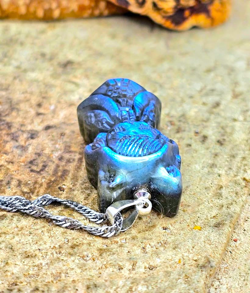 A blue iridescent pendant necklace shaped like a hooded figure, shown close up against a textured brown rock background with part of a silver chain visible in bottom view.-My Aura Gemstones