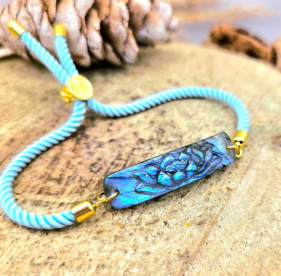 A close-up of a handmade bracelet featuring a light blue and white twisted cord, gold-toned metal accents, and a central rectangular glass bead with a blue lotus flower design.