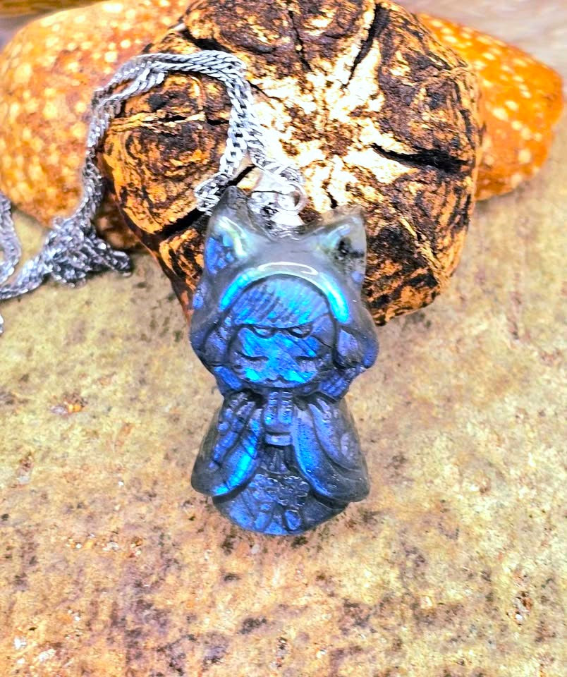 A blue iridescent pendant necklace shaped like a hooded figure, shown close up against a textured brown rock background with part of a silver chain visible.-My Aura Gemstones
