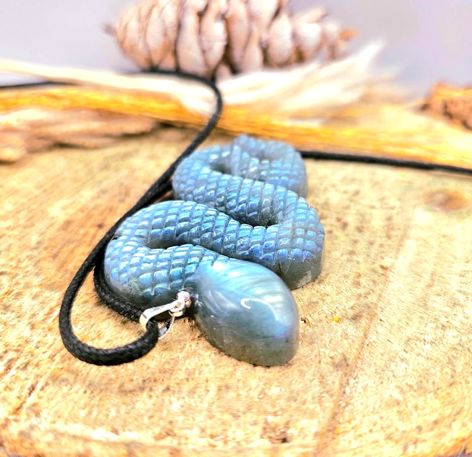 A coiled blue-green labradorite snake pendant necklace on a black cord, displayed on a rustic wood slice with a pinecone in the background.