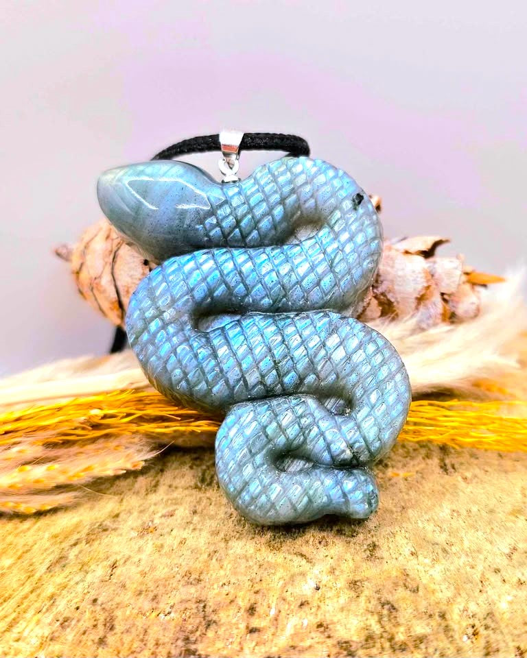 A carved snake pendant with a textured blue-green scale pattern, hanging from a black cord, displayed on a natural wood surface with dried grass and small flowers.