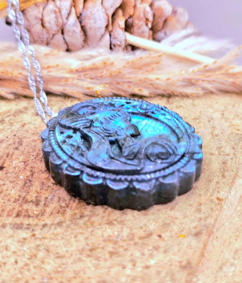A round carved pendant necklace with a silver-toned chain, featuring a dark carved figure with bright turquoise-blue highlights, displayed on a textured tan surface with a pinecone and feather in the background.