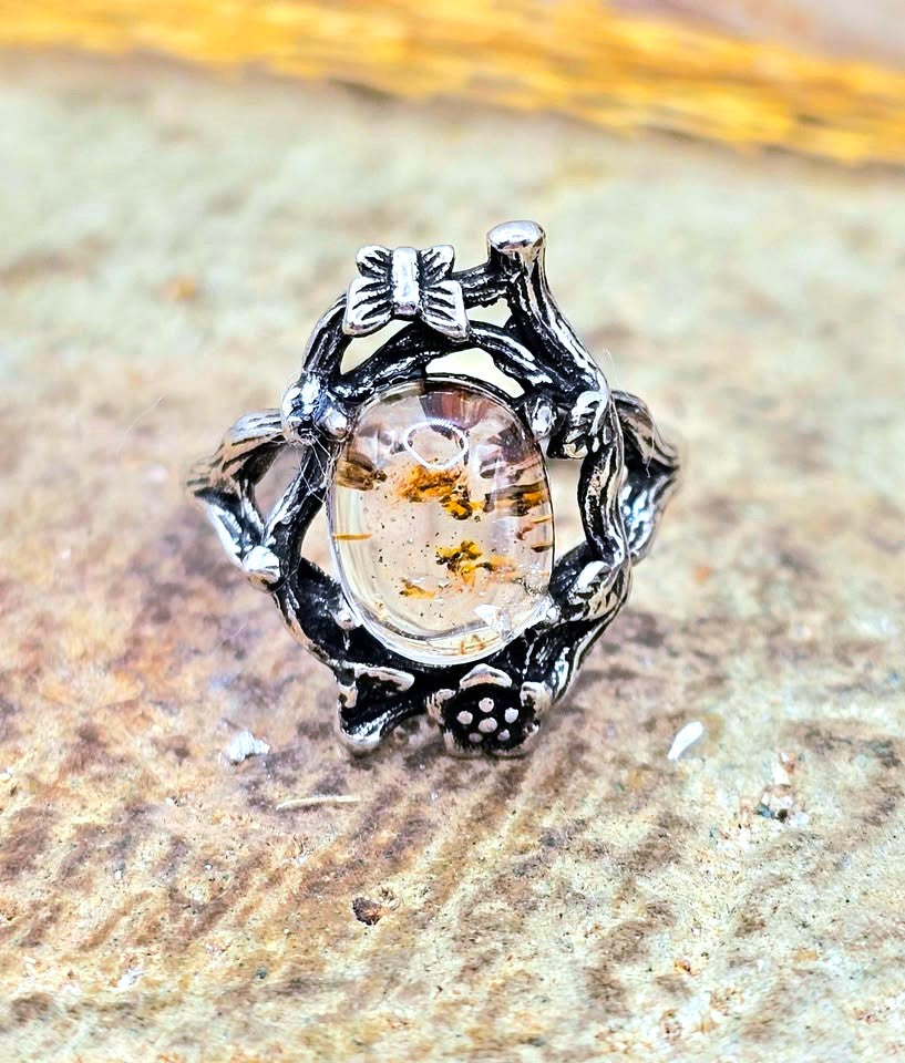 A silver-toned ring with an organic, branch-like sculpted design and a clear oval center containing amber-colored inclusions, resting on a textured stone surface.