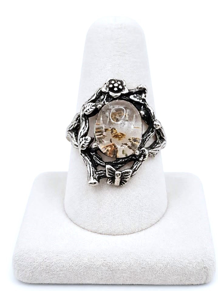 A silver-toned ring with an intricate floral and leafy design surrounding a clear oval cabochon filled with small gold and brown inclusions, displayed on a white ring cone stand against a white background.