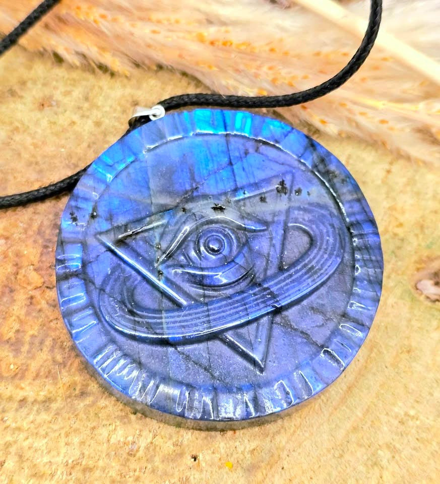 A circular blue labradorite pendant necklace featuring an engraved eye and triangle symbol, resting on a textured tan surface with dried grass.