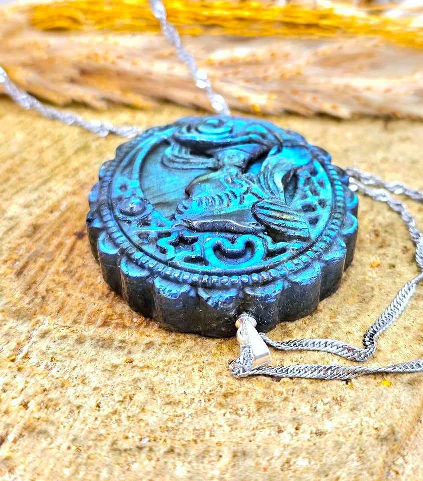 A round carved pendant necklace with a turquoise-blue and black distressed finish, featuring a bird motif in the center, resting on a textured sandy surface with a silver chain.