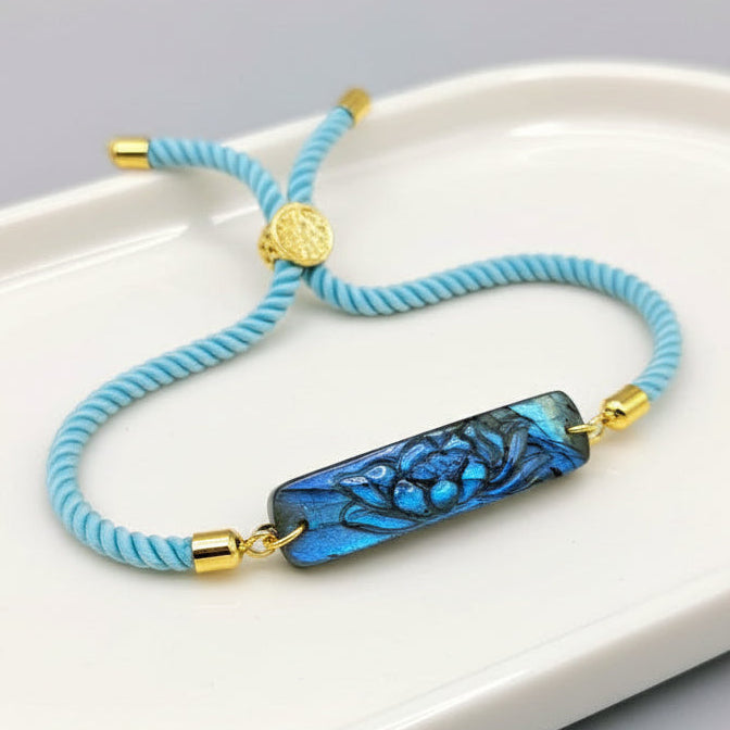 A light blue braided cord bracelet with a polished blue abalone shell pendant featuring a carved fish design, accented with gold-toned metal caps and a small round gold charm, displayed on a rustic wood surface with a pinecone in the background.