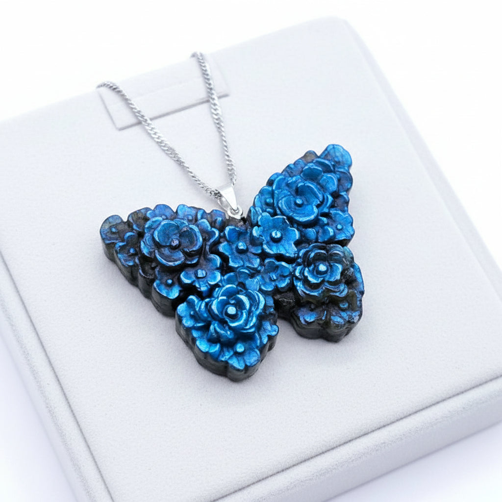 A butterfly-shaped pendant necklace with raised floral details, featuring a vibrant blue and black iridescent finish, hanging from a silver chain against a textured brown background.