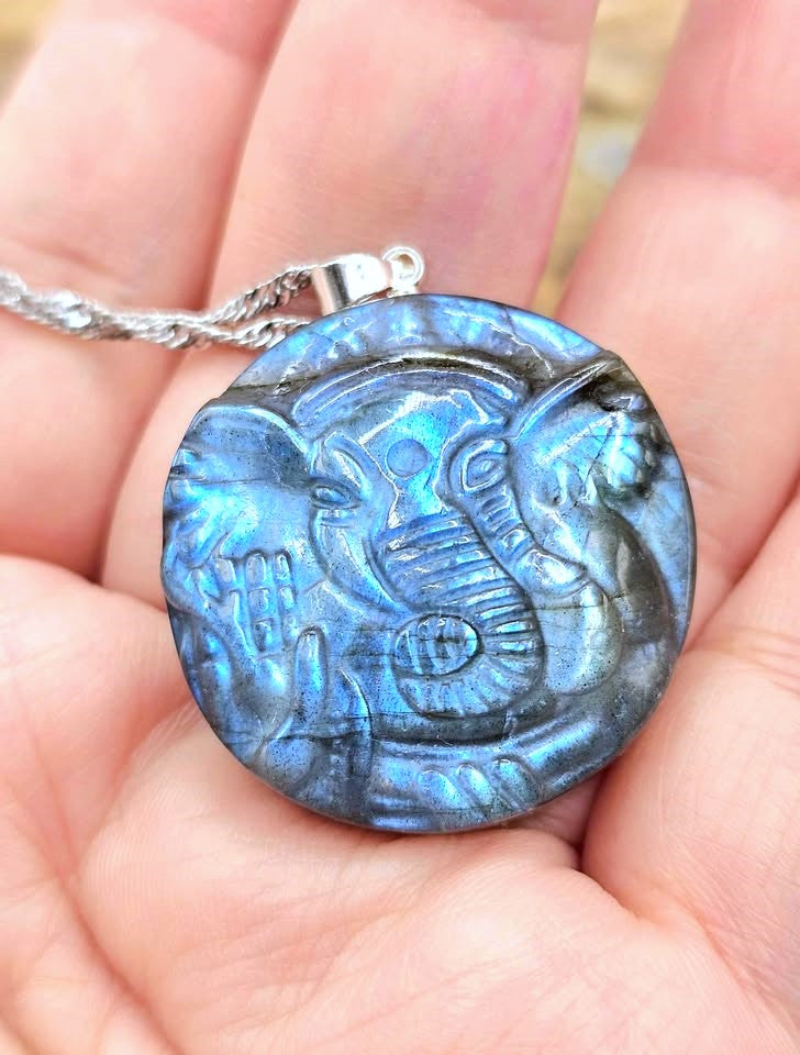 A round labradorite pendant necklace featuring an embossed elephant design, held in a person's hand. The pendant exhibits a shimmering blue and gray iridescent finish, suspended from a silver-toned chain.