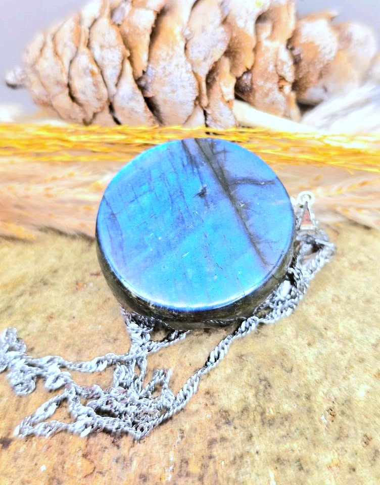 A round labradorite pendant necklace with a silver-toned chain, resting on a textured wooden surface with a pinecone in the background.