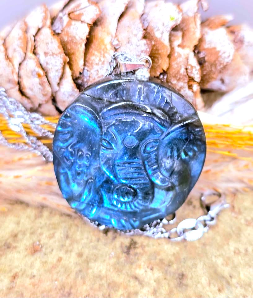 A round blue carved pendant with a silver-toned chain, displayed against a natural background of pinecones and wood.