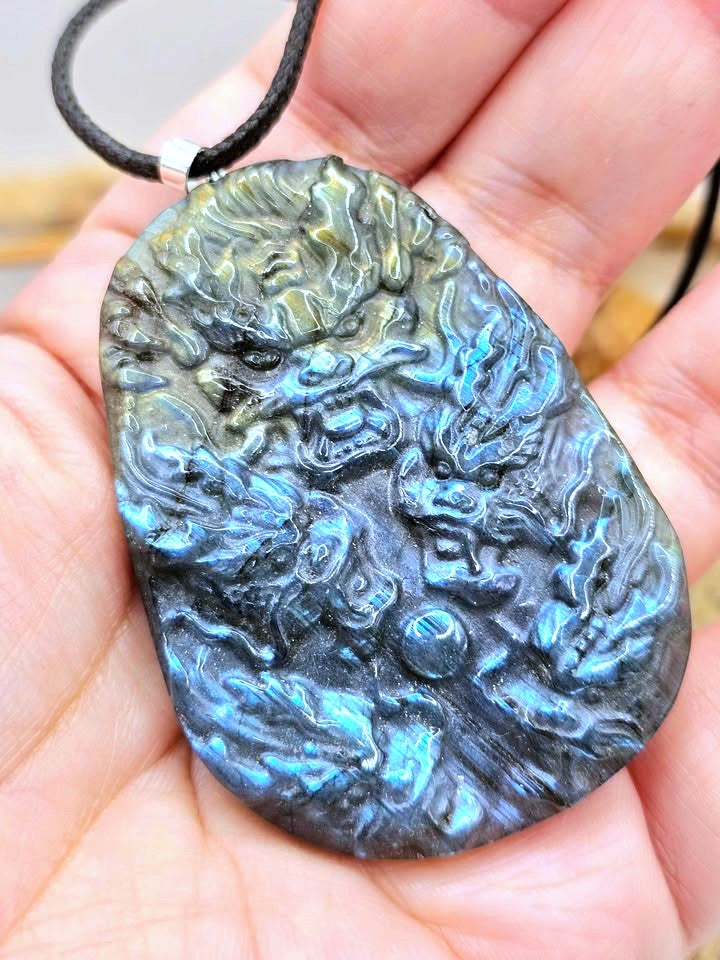 A hand holding a carved labradorite pendant necklace featuring a detailed dragon design. The pendant exhibits a natural blue-green iridescent labradorite flash on a dark gray base, suspended from a black cord.