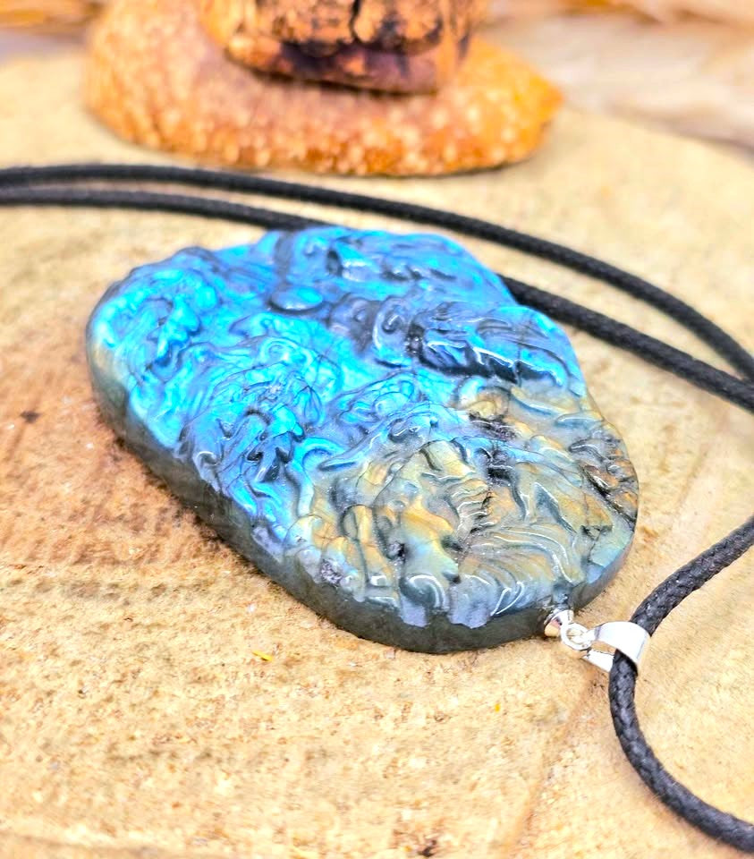 A carved stone pendant necklace with swirling blue, black, and gold marbled patterns, suspended from a black cord on a sandy surface.
