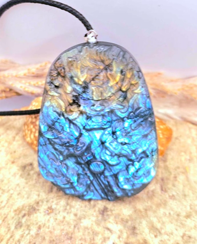 A carved labradorite pendant necklace featuring a detailed dragon design with vibrant blue, green, and gold iridescent flashes, suspended on a black cord over a sandy background.