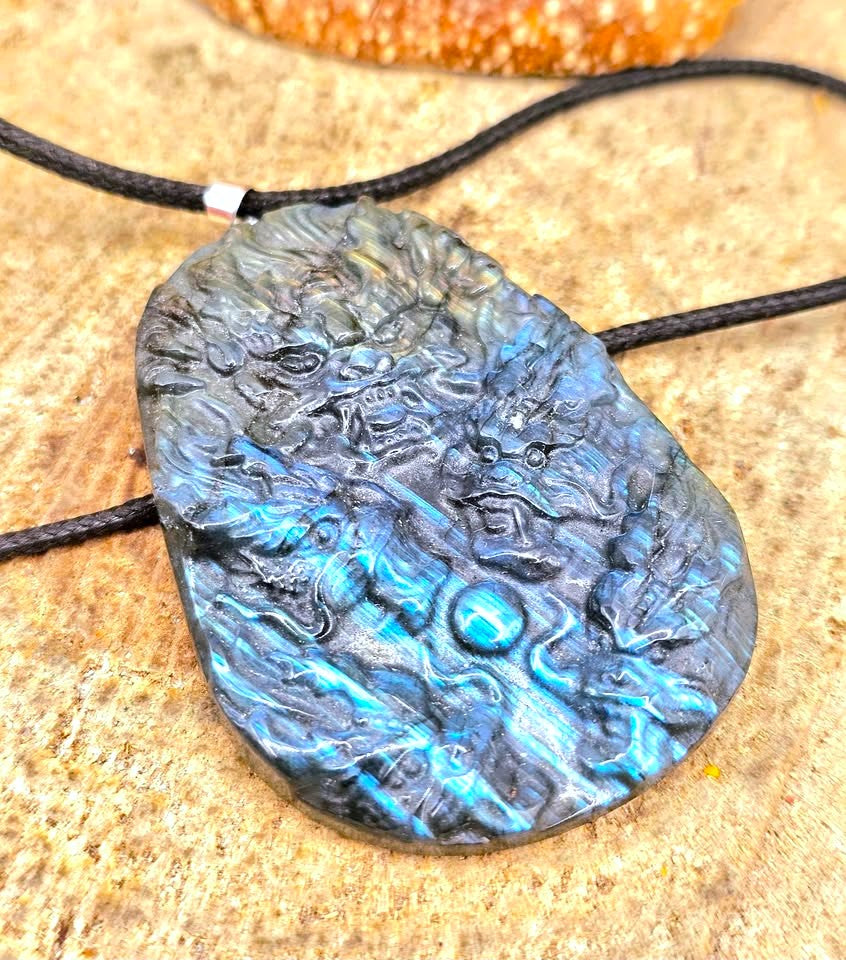 A close-up of a carved black stone pendant with swirling blue and turquoise inclusions, threaded on a black cord, resting on a textured tan surface.