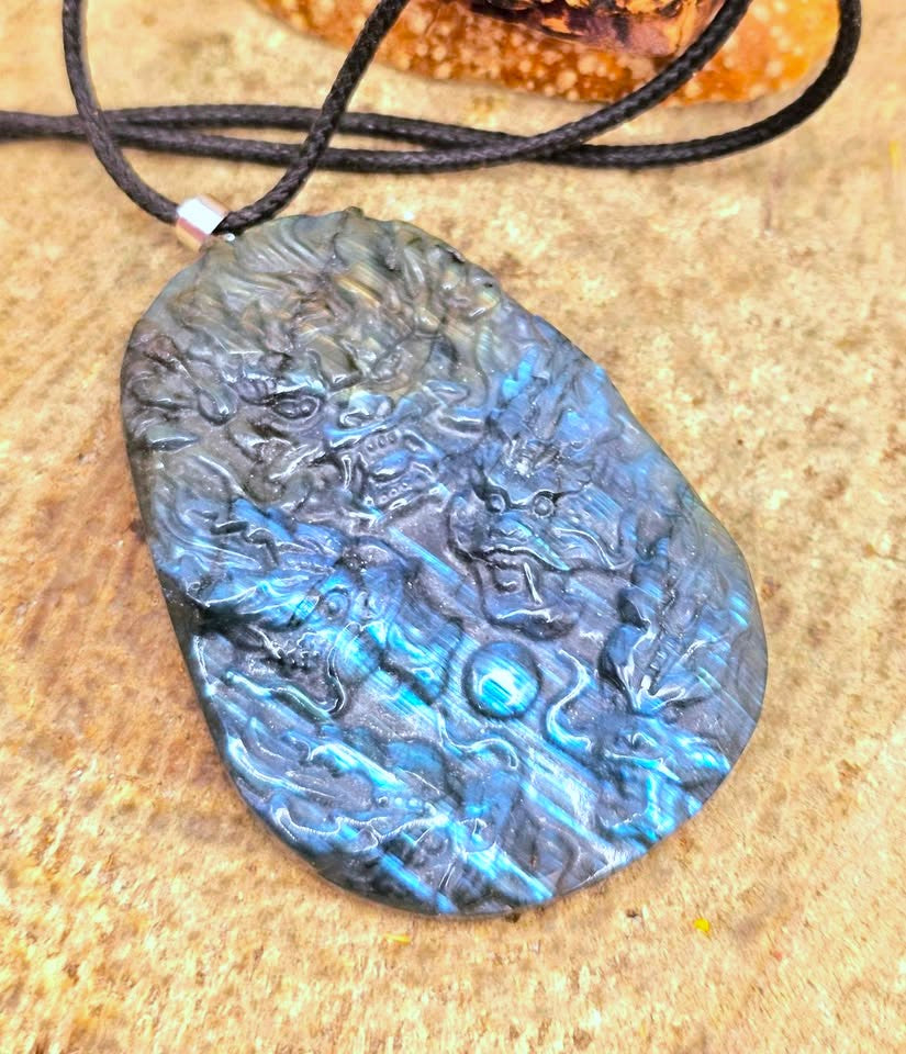 A carved black labradorite pendant necklace with a blue and green iridescent play-of-color effect, hanging on a black cord over a textured tan stone surface.