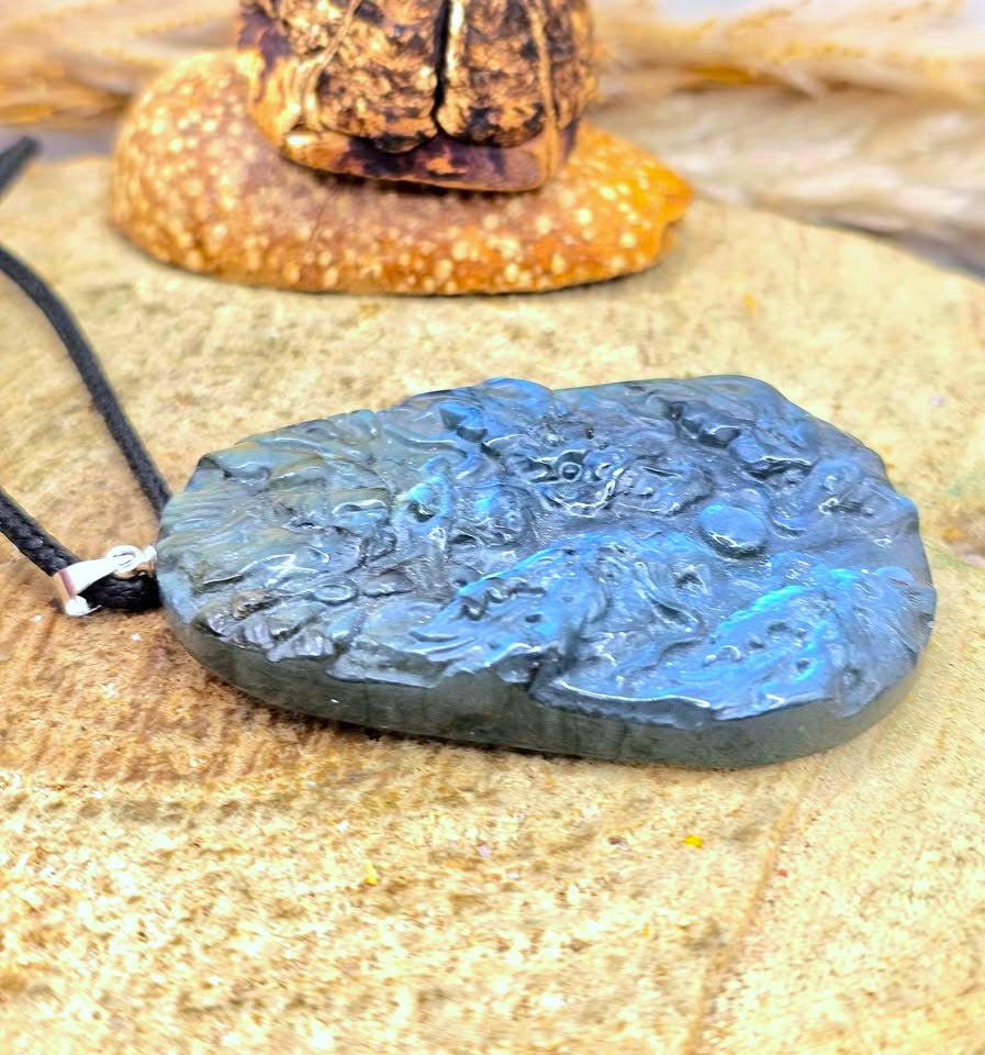 A carved labradorite pendant necklace with a black cord, resting on a sandy surface with a starfish in the background.