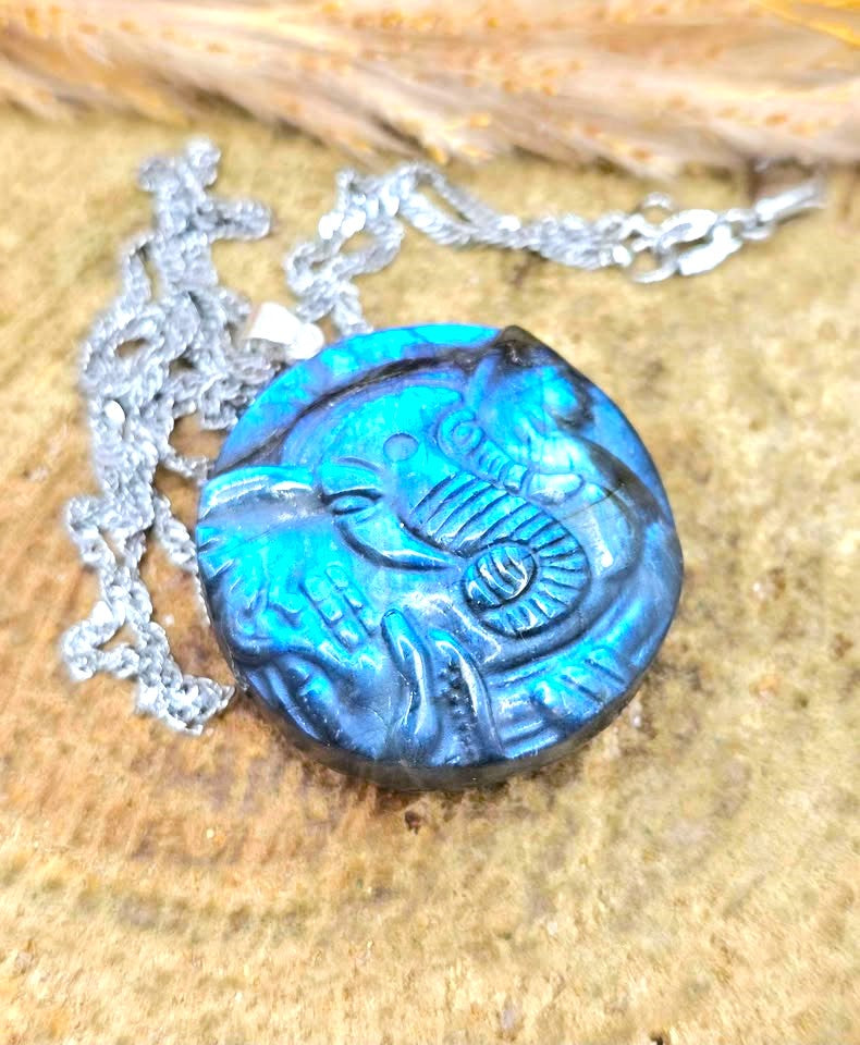 A round carved pendant necklace with a blue and black textured design, resting on a sandy surface with a silver chain.