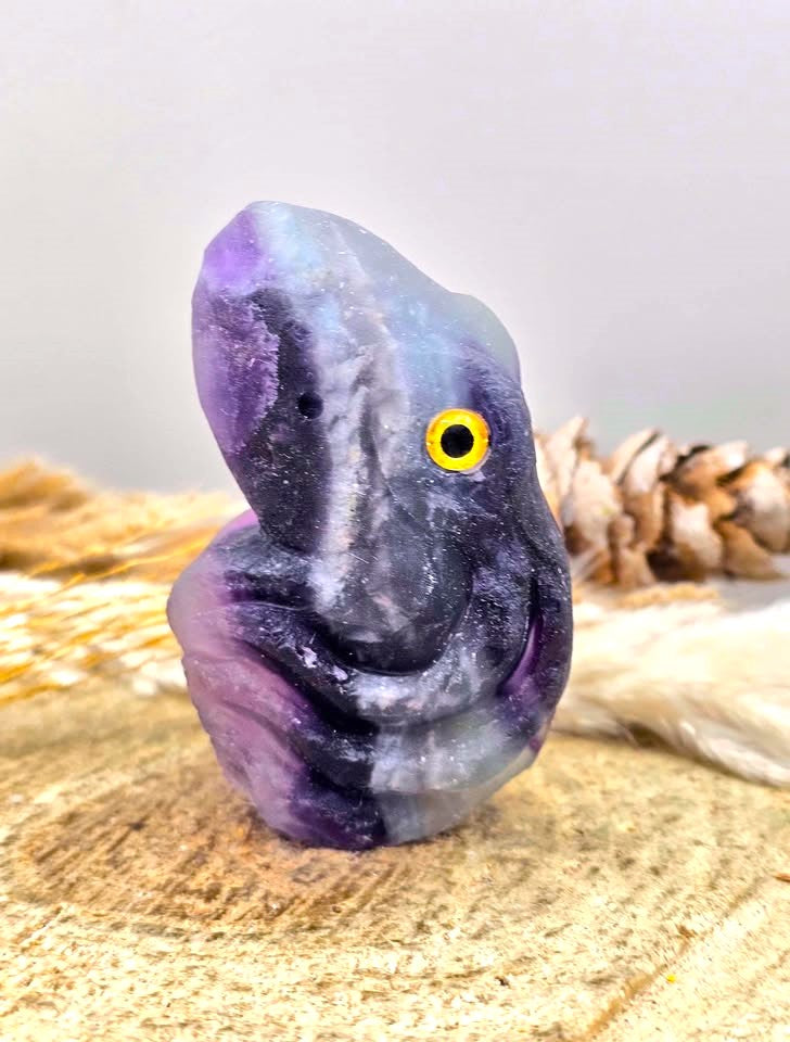A polished purple and blue amethyst crystal figurine shaped like a stylized face with a yellow eye, displayed on a wooden slice with dried grass and a pinecone.