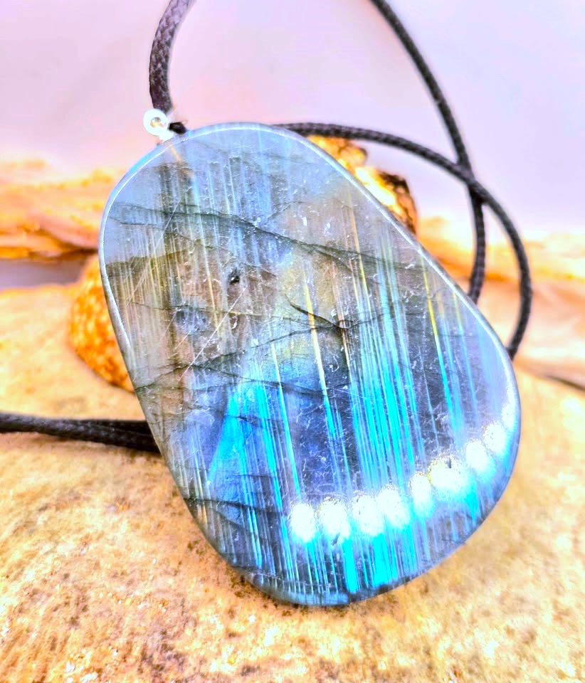 A polished labradorite pendant necklace with a black cord, featuring vibrant blue and green iridescent flashes and fine needle-like inclusions. The pendant is displayed on a textured sandy surface.