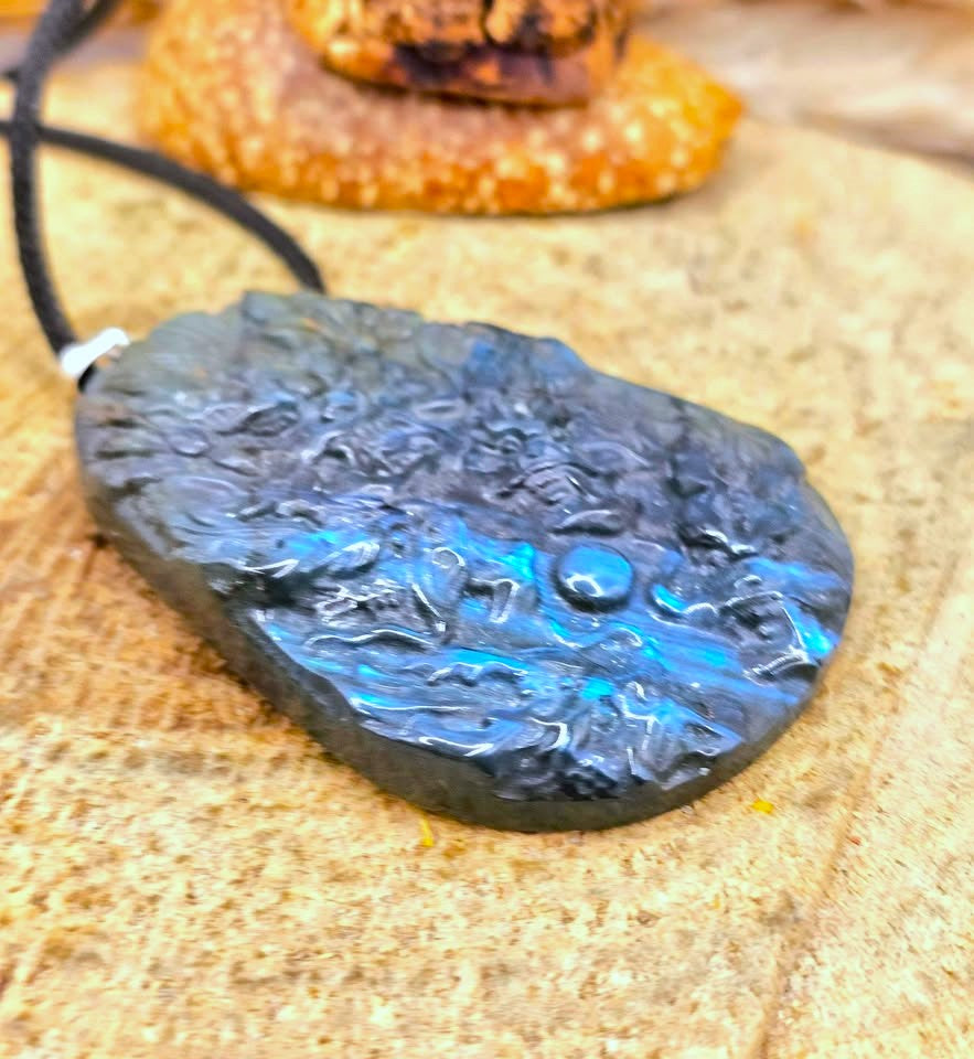 A close-up of a carved black stone pendant with a black cord, featuring a glossy blue iridescent sheen, resting on a textured tan stone surface.