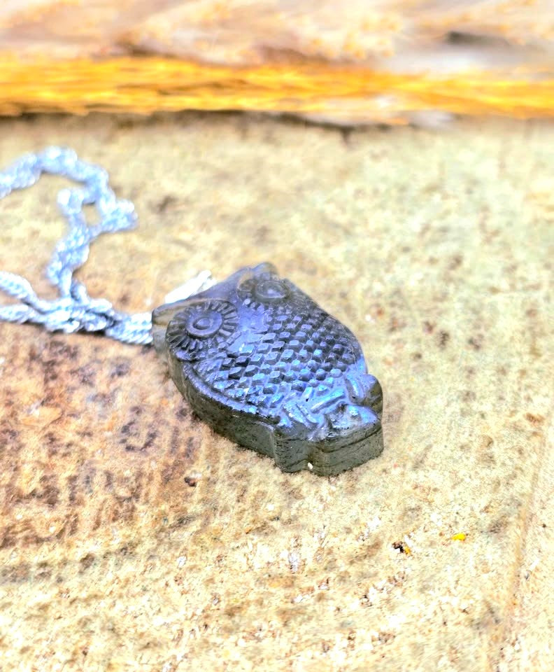 A small owl-shaped pendant necklace with a textured, scale-like pattern in dark gray and metallic blue, resting on a textured stone surface with a silver chain partially visible.