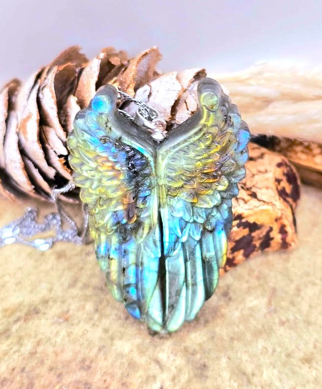 A carved angel wing pendant with a blue, green, and gold iridescent finish, resting on a pine cone and textured surface.