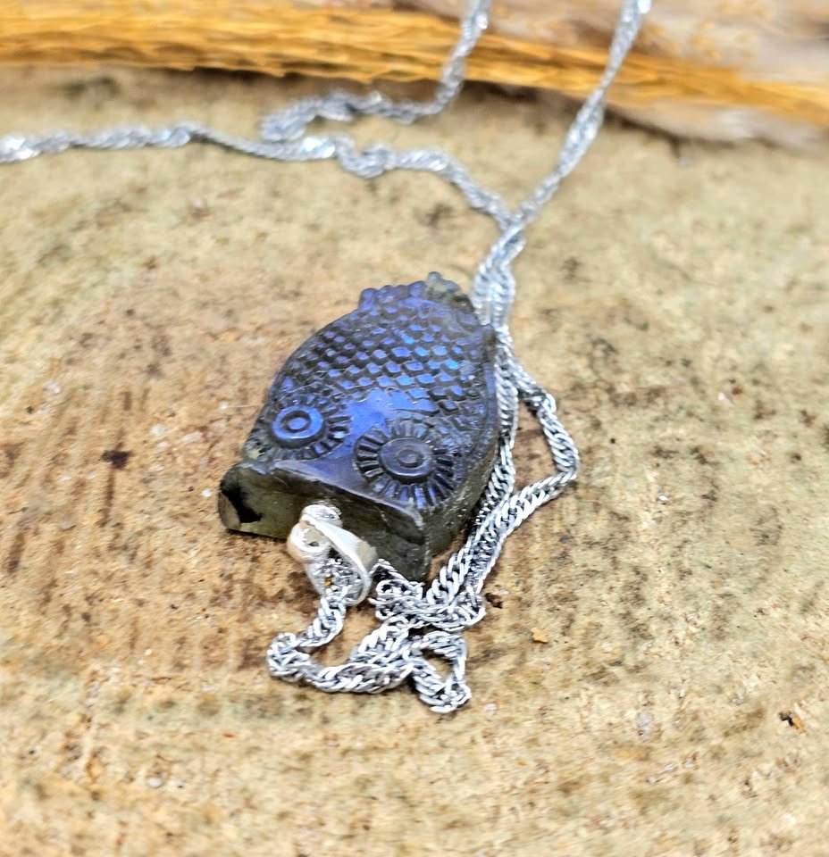 A silver chain necklace with an owl-shaped pendant made from dark stone or resin, photographed close up on a textured tan surface.