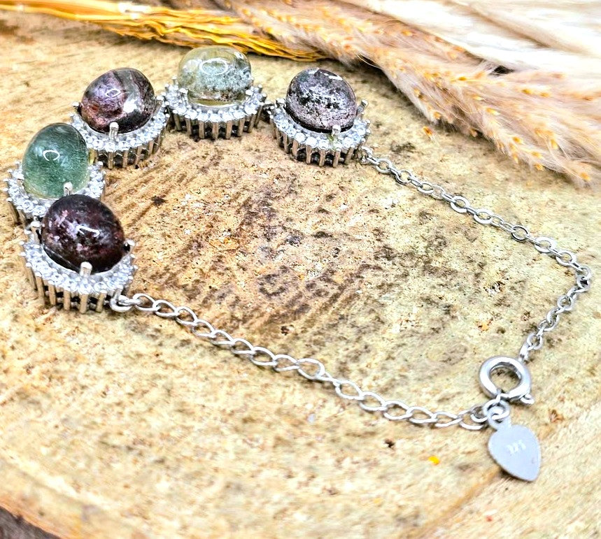 A silver chain bracelet with five round gemstone cabochons in silver-toned settings, displayed on a textured wood surface with dried grass in the background.