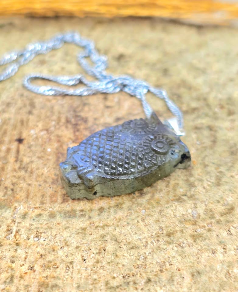 A silver fish-shaped pendant necklace with a textured scale pattern, resting on a sandy, natural background.