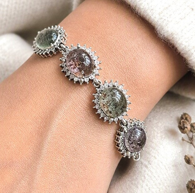 A close-up of a silver-toned bracelet with three oval multicolor gemstones and small clear stones, worn on a person's wrist with a cream knit sleeve visible.