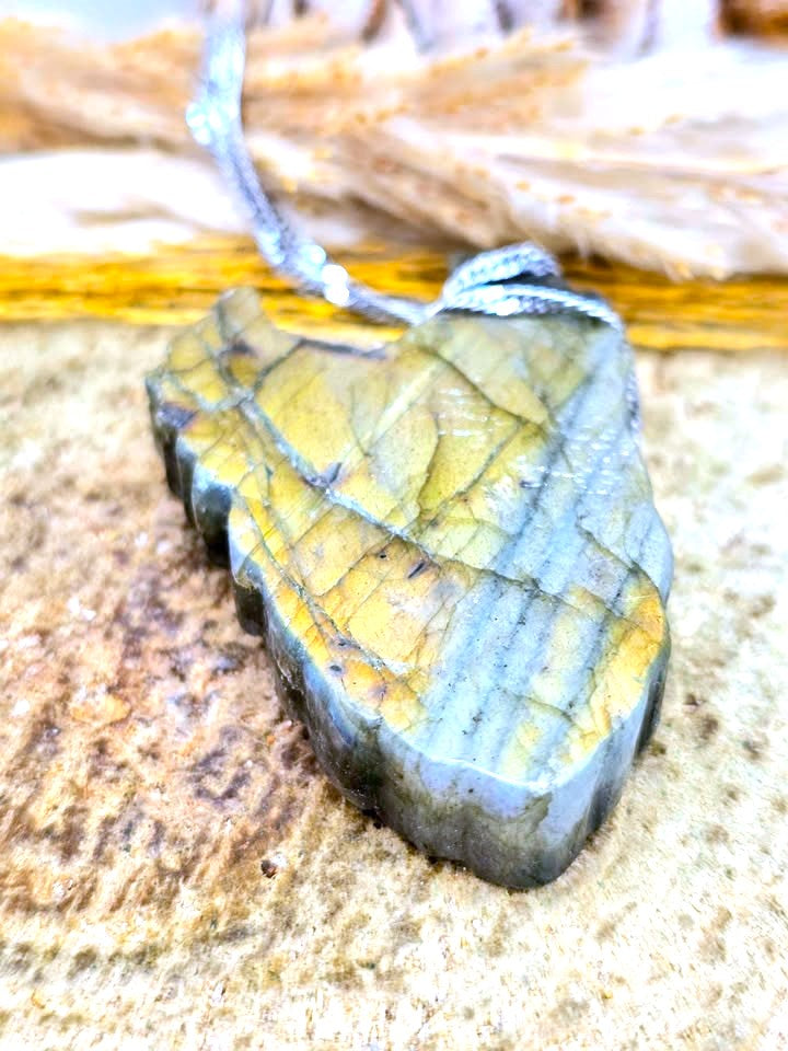 A raw labradorite stone pendant with a silver bail and cord, resting on a textured stone surface.