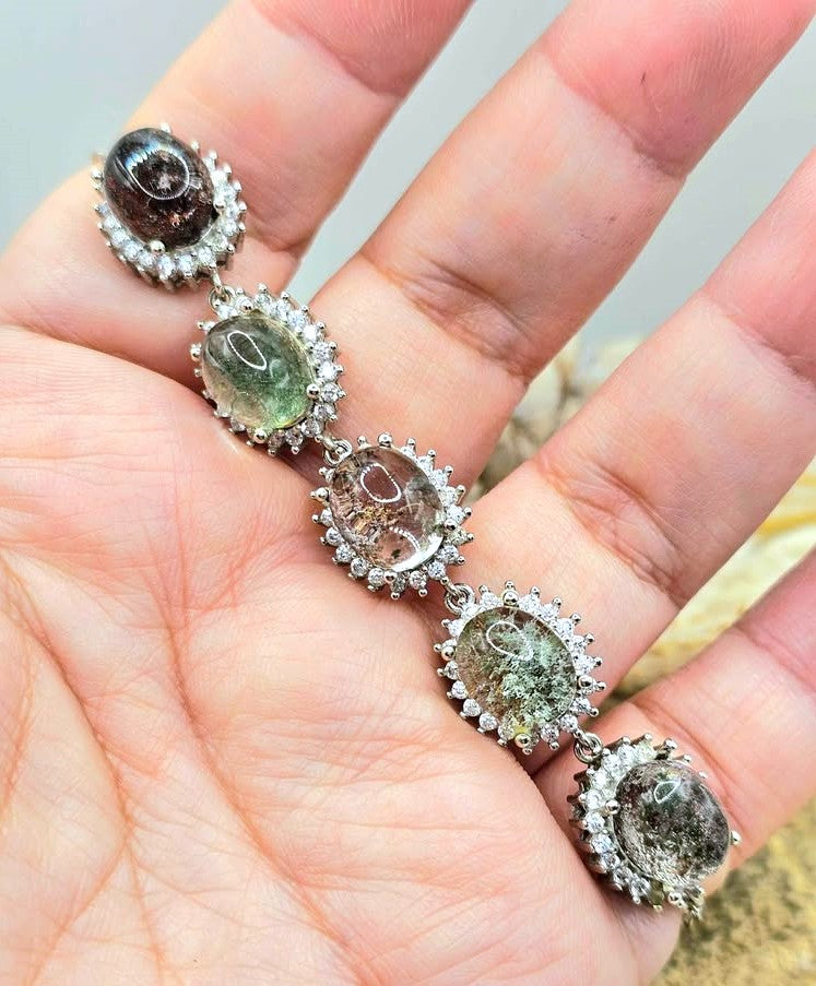 A close-up of a hand displaying a silver-toned link design bracelet with five oval clear gemstones, each surrounded by small clear stones and set with a larger greenish stone above.