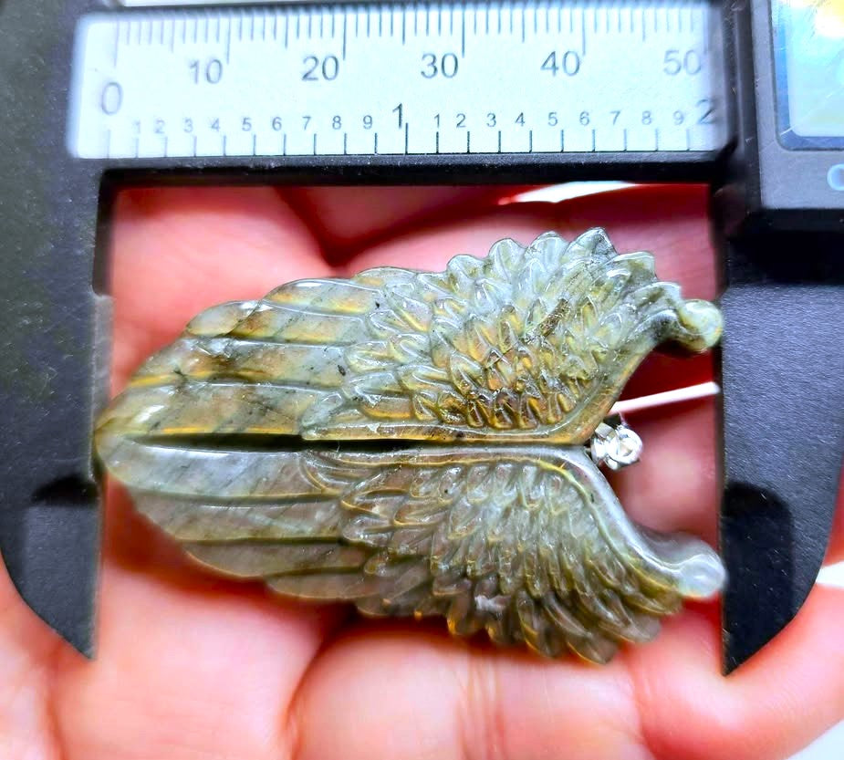 A close-up of a carved wing-shaped brooch or pendant with a small clear gemstone, held in a hand under a black caliper for size reference. The item has a greenish-gold marbled color with darker shading.