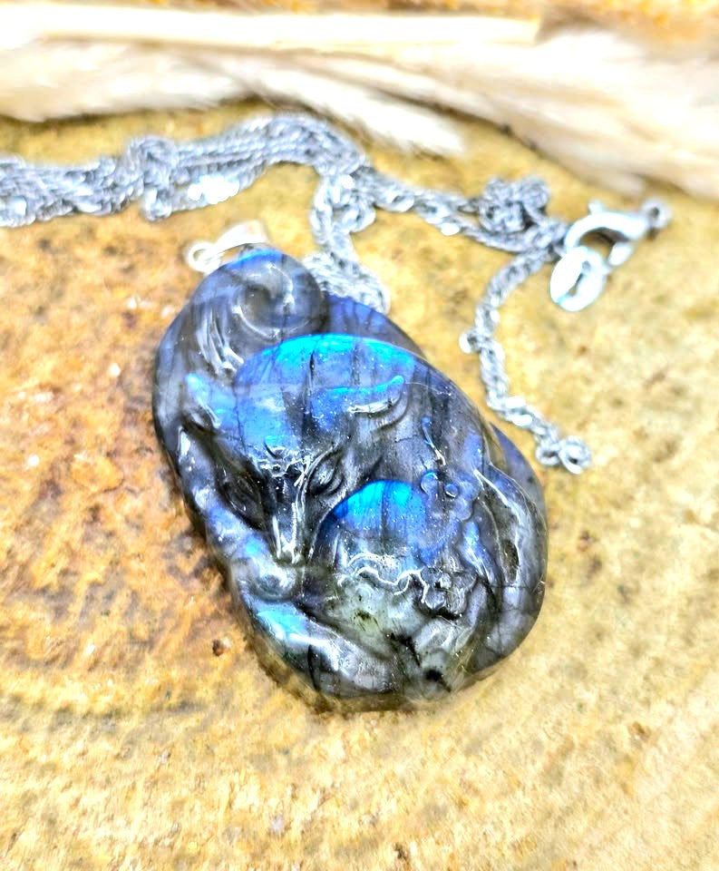 A carved labradorite pendant necklace featuring a detailed fox design with vibrant blue and green iridescent flashes, resting on a textured tan stone surface with a silver chain.