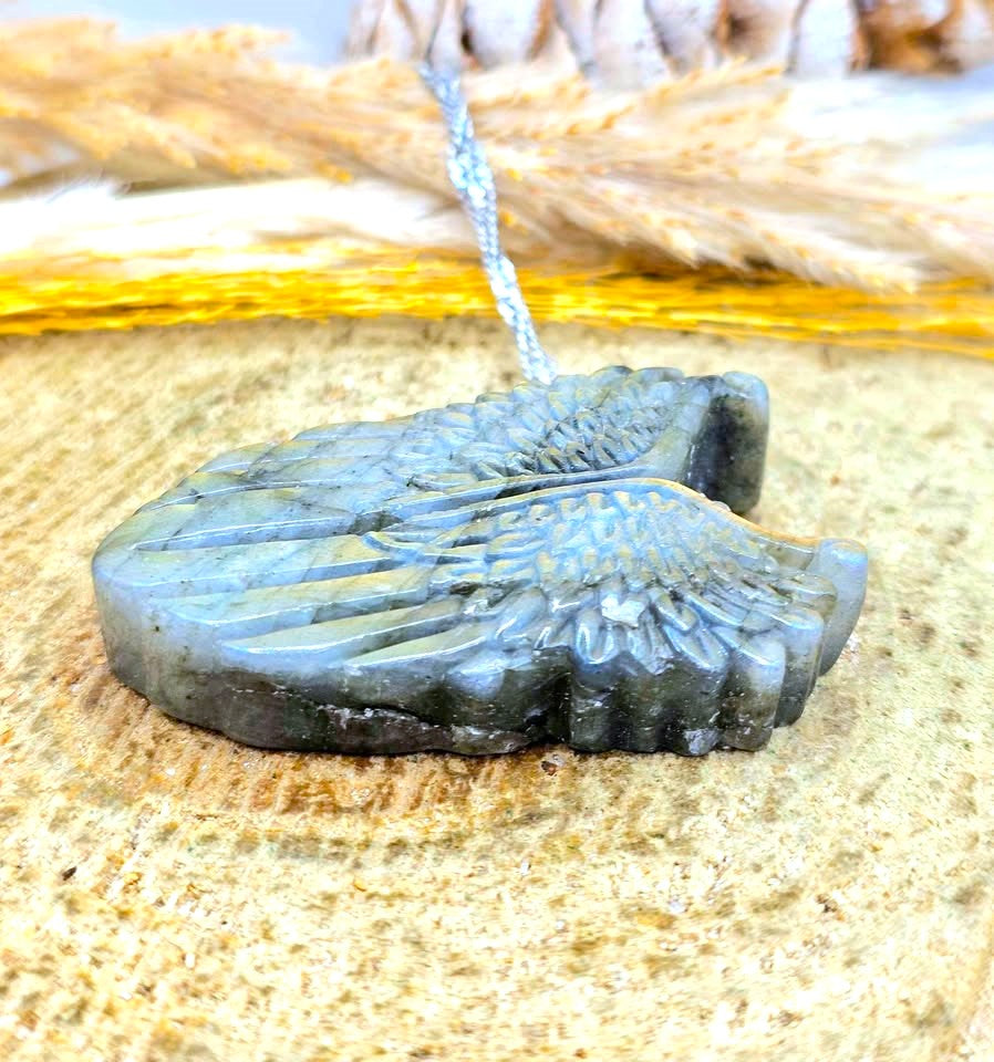 A carved Labradorite pendant shaped like a pair of wings, resting on a textured beige surface with dried grass and gold foil in the background.