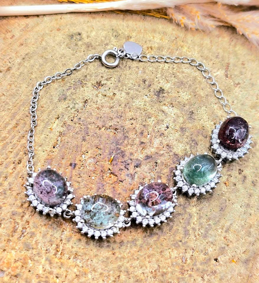 A silver chain bracelet featuring five oval multicolored gemstones, each surrounded by a small halo of clear stones, displayed on a textured brown stone surface.
