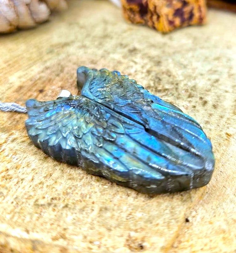 A carved labradorite wing pendant with shimmering blue and green iridescence, resting on a textured tan stone surface.