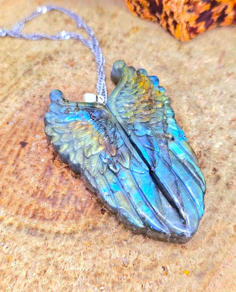 A wing-shaped pendant necklace with a textured, layered design in shades of blue, green, and gold, hanging from a silver cord on a rustic wood surface.