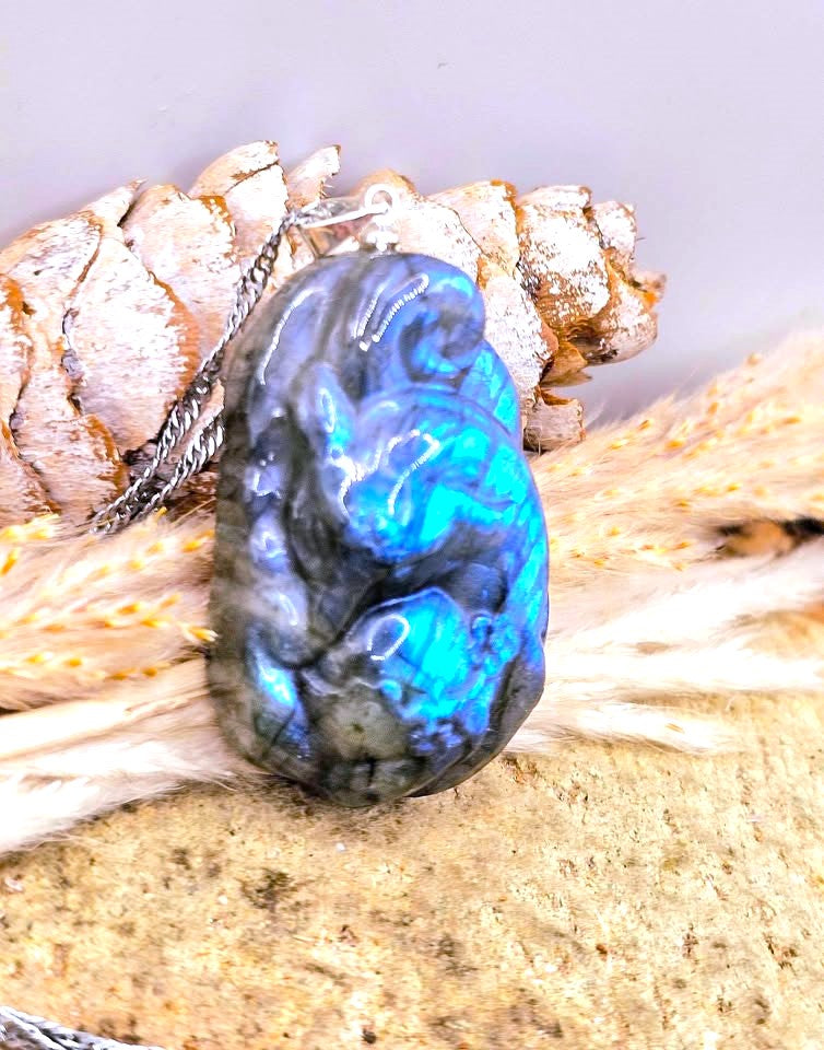 A polished labradorite stone pendant with vibrant blue and green iridescent flashes, hanging from a silver chain and resting on dried grass and a pine cone.