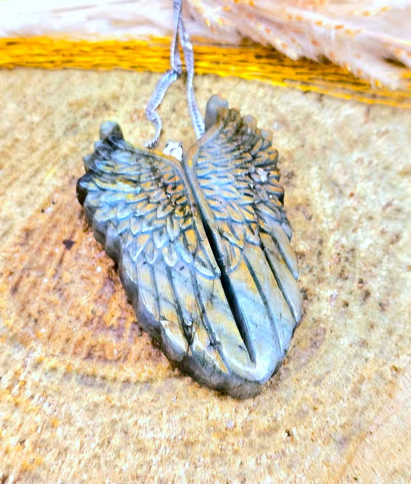 A carved stone angel wing pendant with a gray stone body and gold-toned highlights, hanging from a silver cord against a textured tan background.