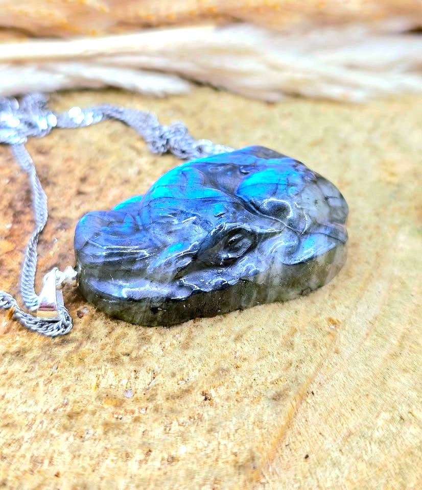 A raw, polished Labradorite pendant necklace with a silver chain, resting on a textured sandy surface. The pendant displays vibrant flashes of blue and green iridescence over a dark gray base.