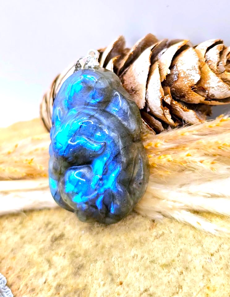 A polished labradorite pendant with vibrant blue and green iridescent flashes, resting on a textured beige surface next to a pinecone and dried grass.
