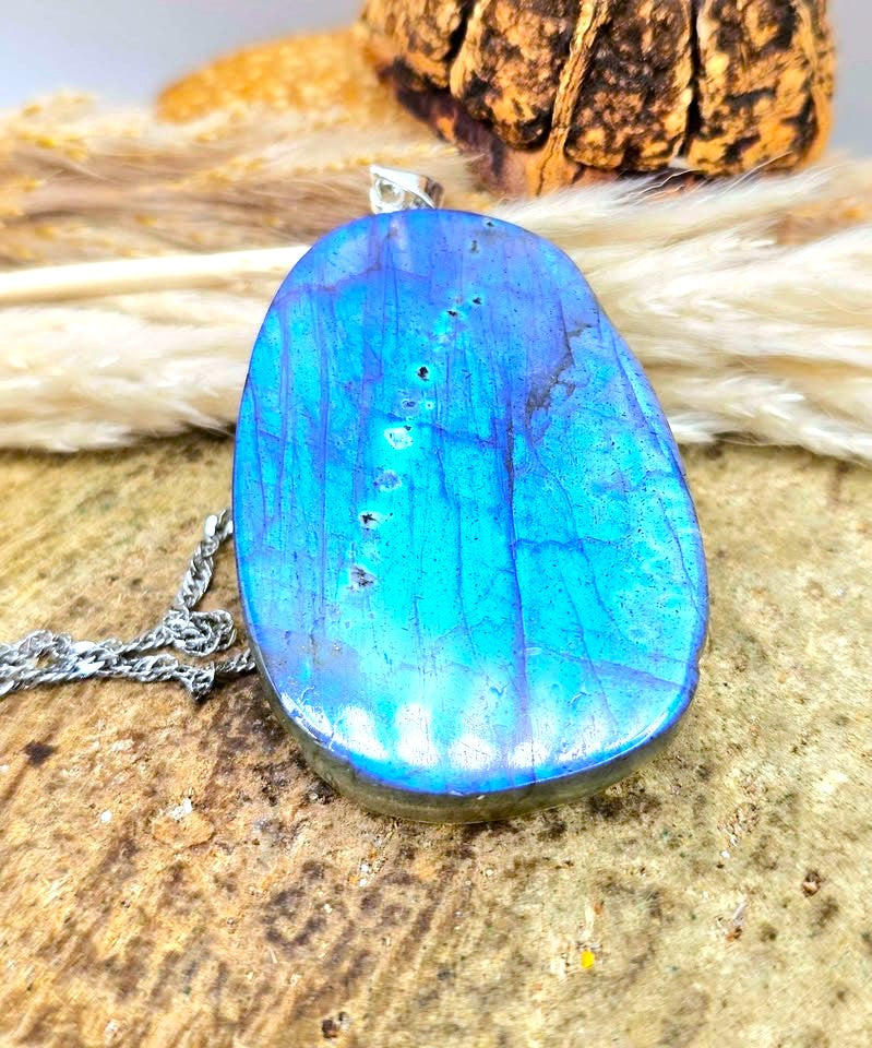A polished blue labradorite pendant necklace with a silver bail and chain, displayed on a textured stone surface with dried grass and a piece of wood in the background.