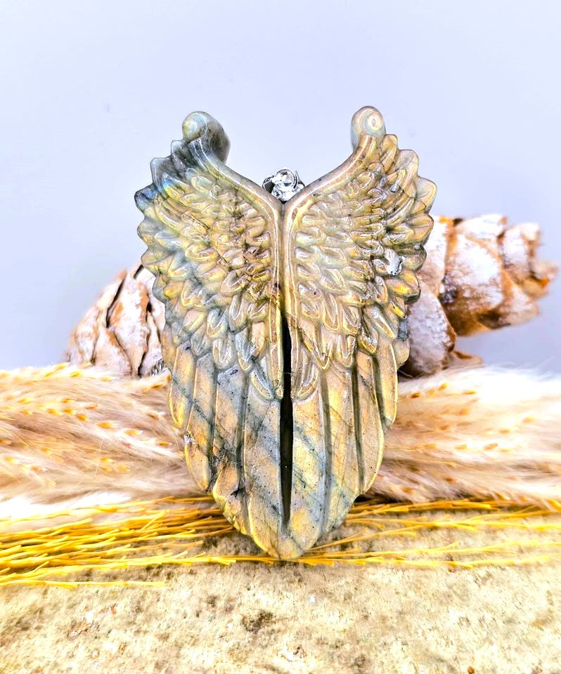 An angel wing ring with a textured, layered finish in gold, bronze, and gray tones, displayed on a rustic background of dried grass and pine cones.