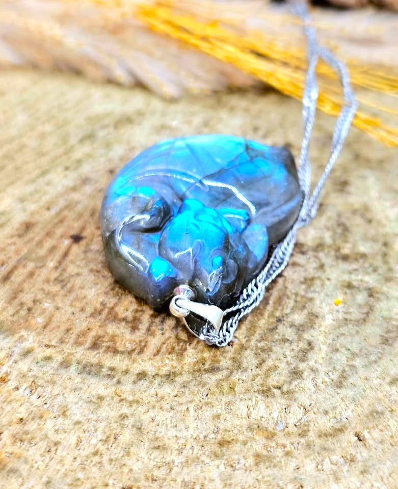 A close-up of a polished blue and black opal pendant necklace on a silver chain, resting on a textured tan stone surface.