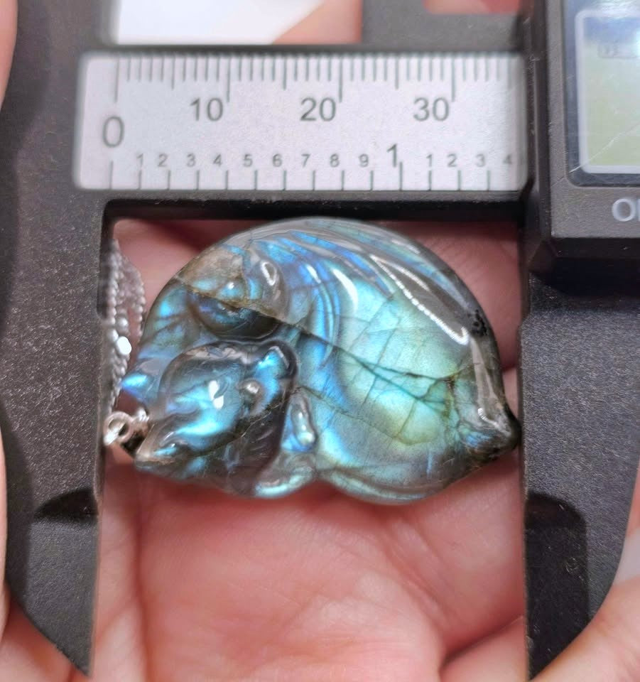 A polished labradorite pendant with a blue-green iridescent flash, held in a hand under a digital caliper to show its dimensions.
