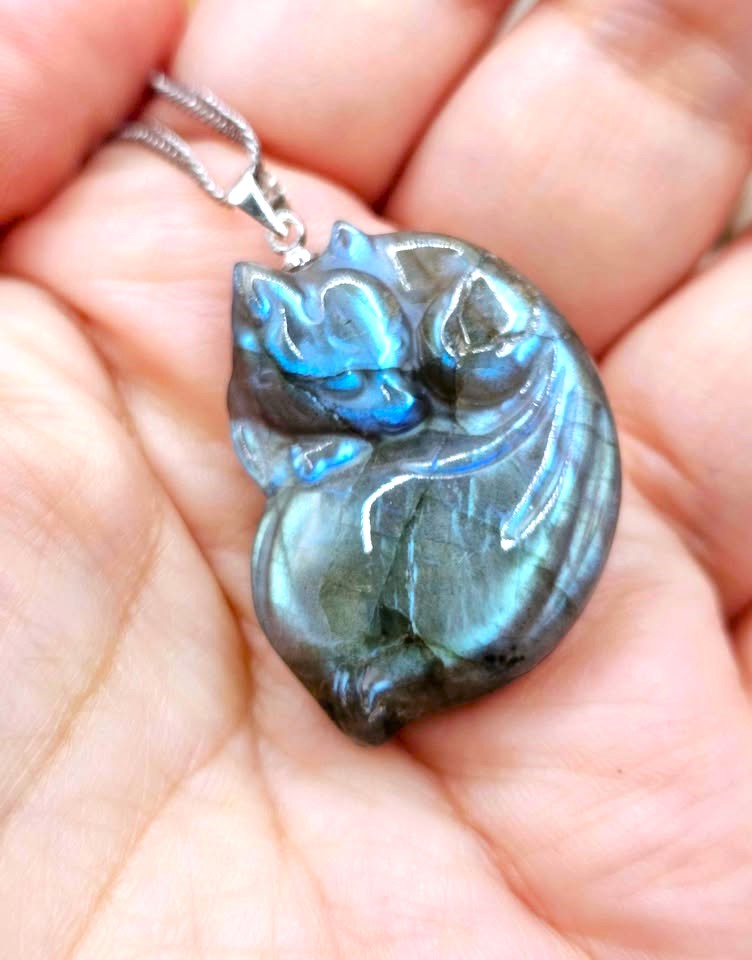 A close-up of a hand holding a polished labradorite pendant shaped like a sleeping cat on a silver chain.