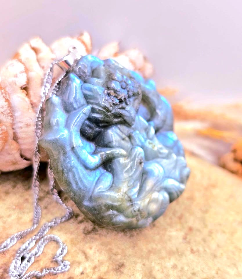 A carved blue-green stone pendant with a silver chain, displayed on a sandy surface next to a seashell.